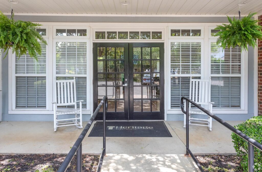 Entrance to Heritage apartments clubhouse with large windows and window-paned door and two outdoor rocking chairs