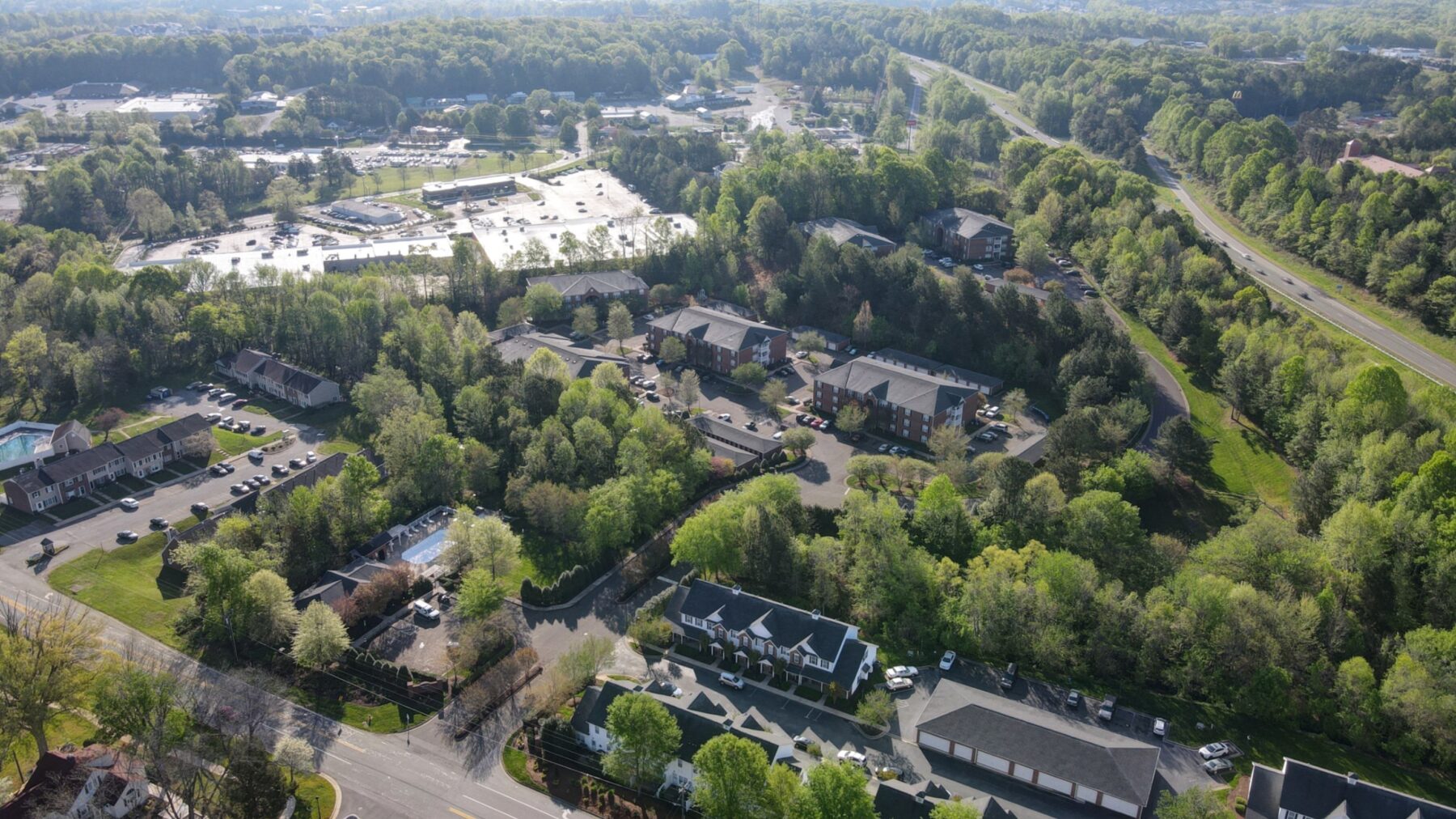 Aerial view of a suburban neighborhood with apartment complexes, tree-lined roads, and a nearby shopping center surrounded by lush greenery.