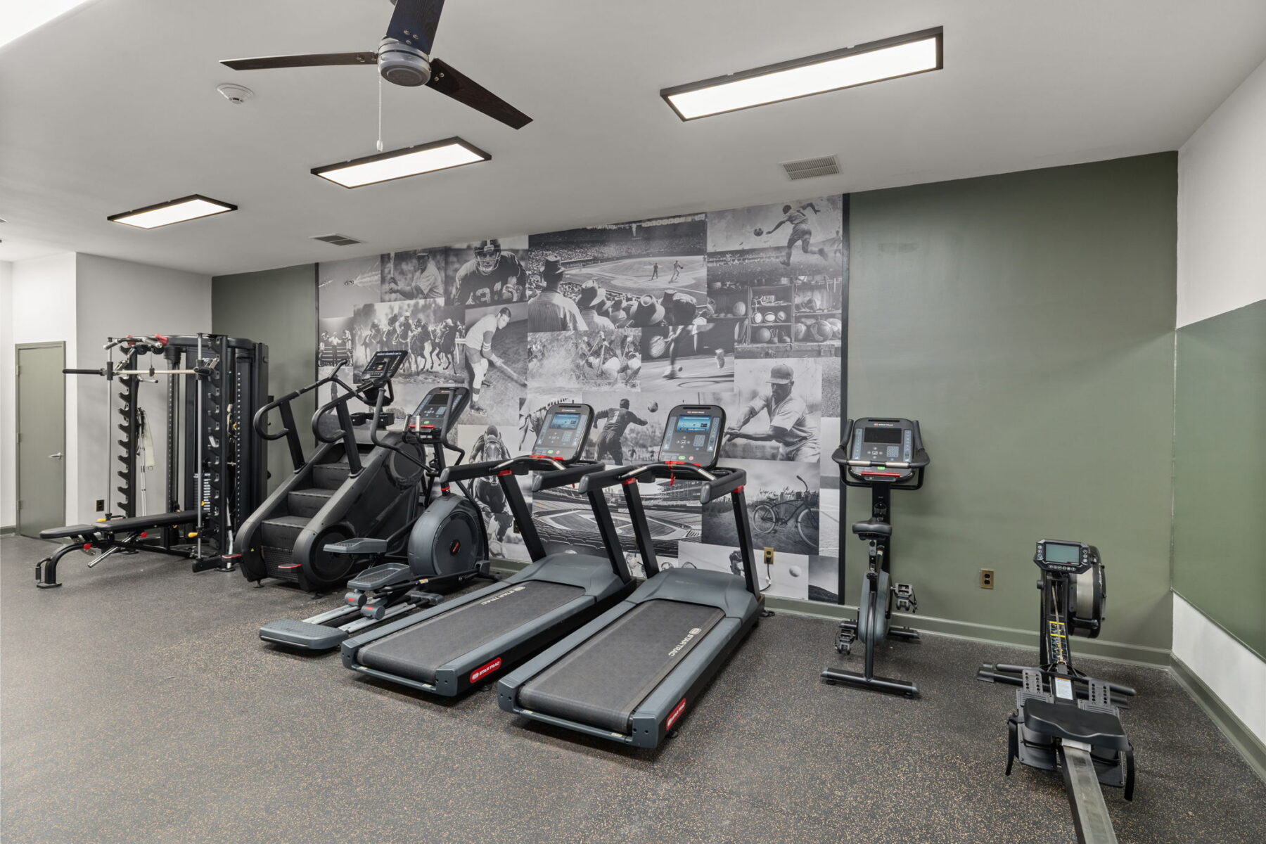 A modern gym room with exercise machines, including two treadmills, an elliptical, a rowing machine, and a multifunctional weight machine. The back wall features a large sports-themed mural.