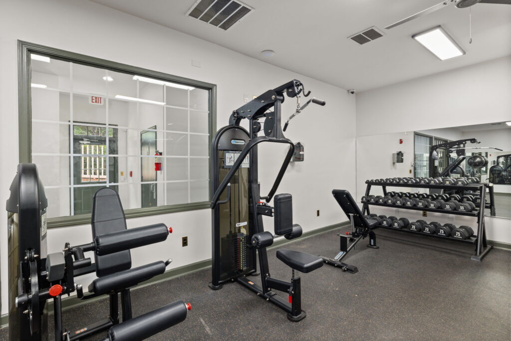 A modern gym room with weight machines, a bench, a rack of dumbbells, a large wall mirror, and a glass window looking into another room. The space is clean and well-lit with white walls and ceiling lights.