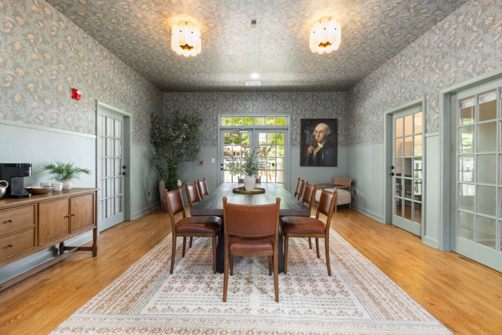 A dining room with floral wallpaper, a long table surrounded by brown leather chairs, wooden floors, a sideboard with a coffee maker, glass doors, large windows, and a portrait of a man on the wall.