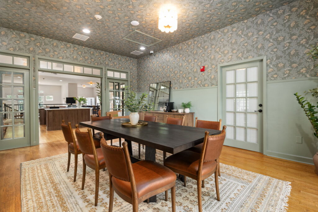 A dining room with a dark wooden table, six brown leather chairs, a patterned rug, and floral wallpaper. Green-trimmed glass doors and windows let in light; plants and decorative items are on the sideboard.