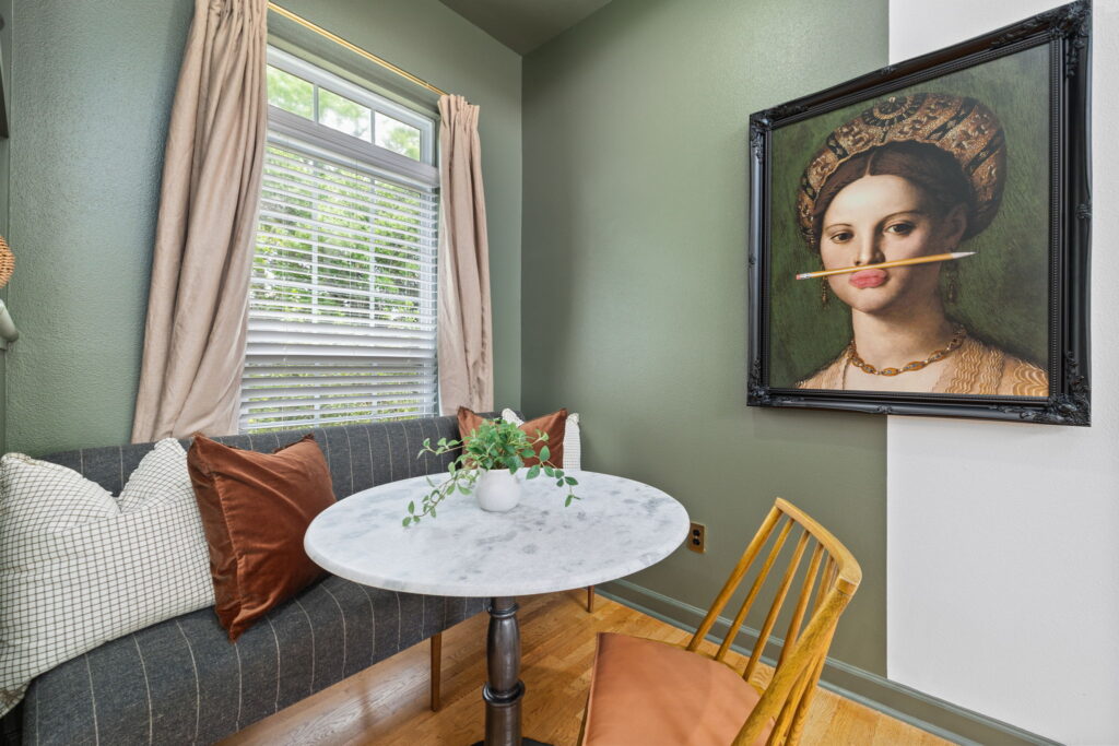 A cozy dining nook with a round marble table, a small plant centerpiece, a cushioned bench with pillows, a wooden chair, and a framed painting of a woman balancing a pencil in her mouth on a green wall.