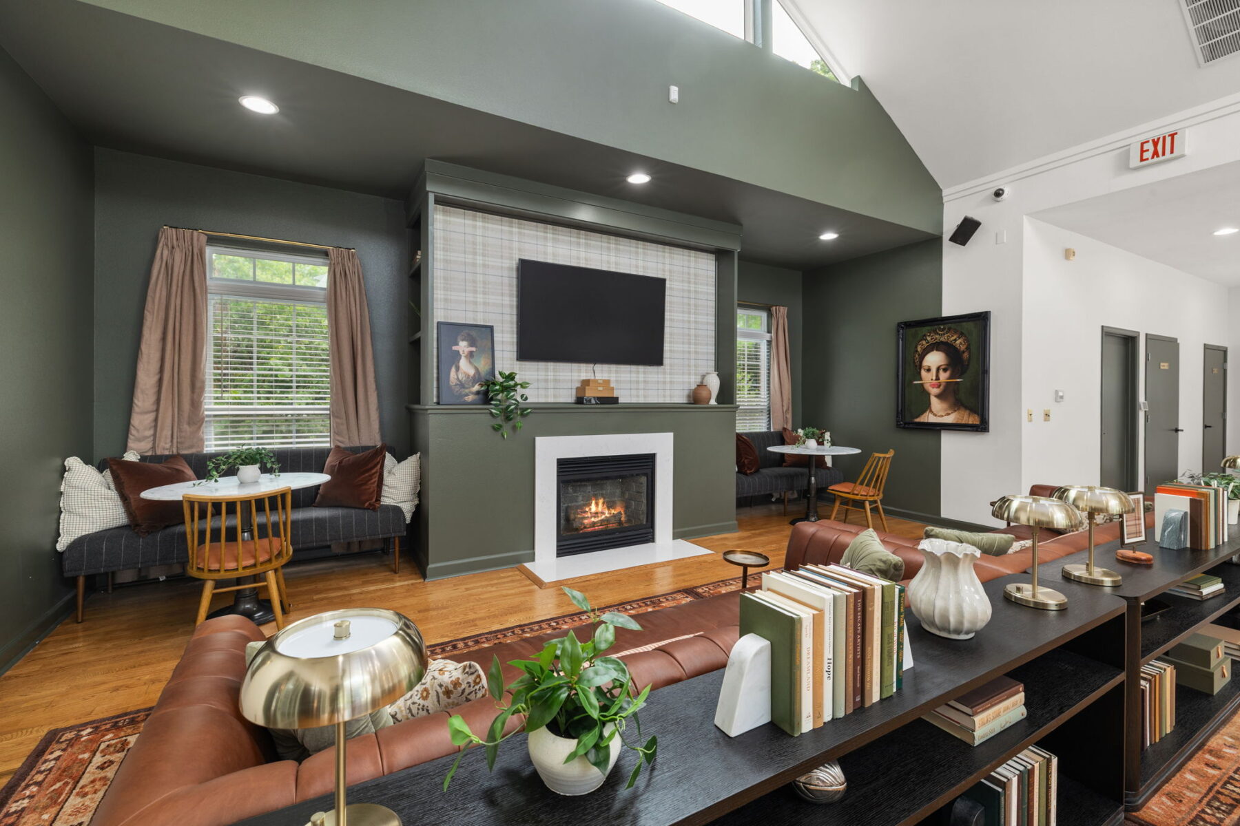 A modern lounge with green walls, a fireplace under a wall-mounted TV, cozy seating areas, bookshelves with decor, and large windows letting in natural light. Artwork and plants add warmth to the inviting space.