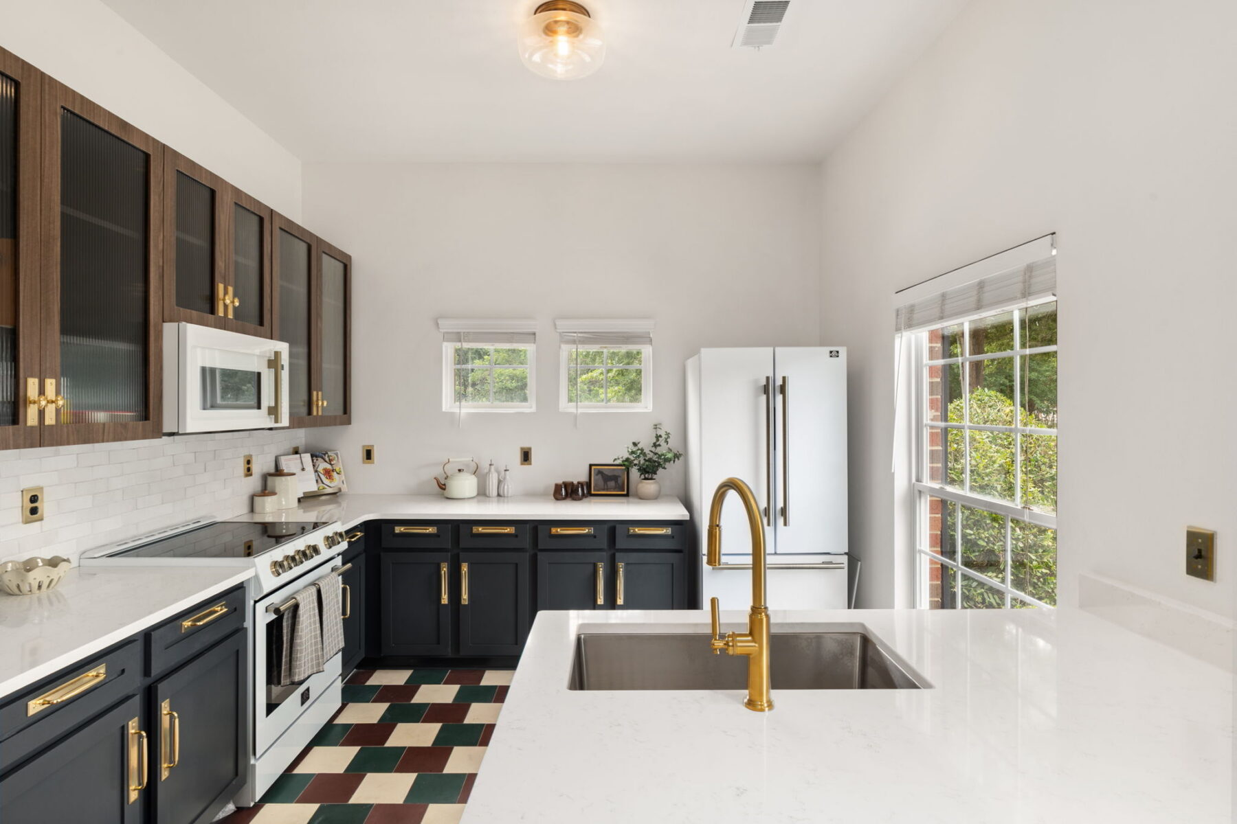 Modern kitchen with white countertops, navy cabinets, brass hardware, and appliances including a stove, microwave, and French door refrigerator. Large windows let in natural light, highlighting the checkered floor.