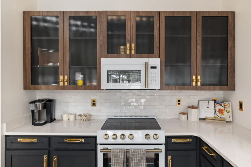 Modern kitchen with dark lower cabinets, white countertop, white stove, and microwave. Upper cabinets have glass doors. Coffee maker, cups, and cooking utensils sit on the counter; towel hangs on oven handle.