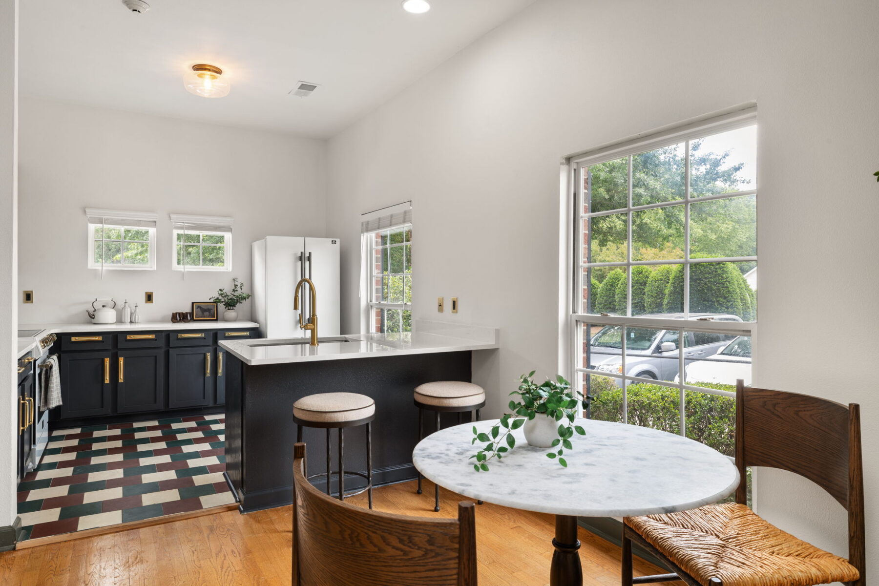 Bright kitchen and dining area in apartments in Hillsborough, NC, featuring black cabinets, white countertops, a gold faucet, patterned tile floor, and a round marble table with wooden chairs by a large window overlooking greenery and a parked car.