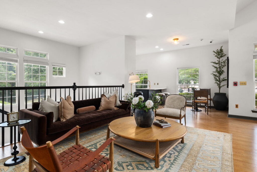 A bright, modern living room with a dark sofa, woven chairs, round wooden coffee table, potted plants, large windows, and light wood flooring. Natural light fills the space, creating a warm and inviting atmosphere.