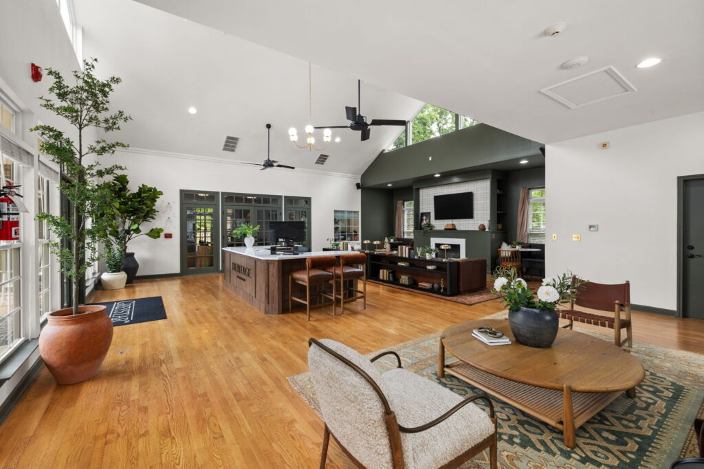 A spacious, modern living area with wood flooring, large windows, a central kitchen island, comfortable seating, a wall-mounted TV above a fireplace, and plants adding greenery to the bright space.