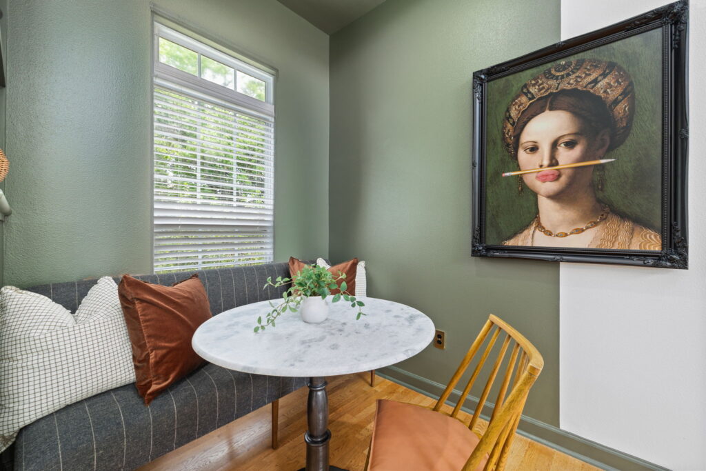 A cozy corner features a small round marble table with a wooden chair and a cushioned bench. On the wall hangs a portrait of a woman humorously depicted with a pencil held in her mouth. A vase with greenery sits on the table.