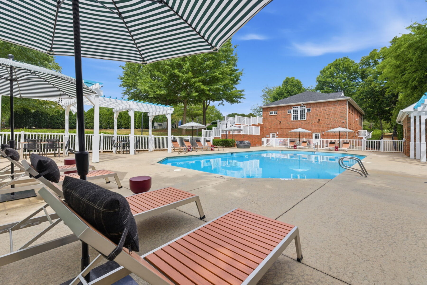 Outdoor swimming pool surrounded by lounge chairs, striped umbrellas, and white pergolas at apartments in Hillsborough NC, with a brick building and trees in the background under a blue sky.