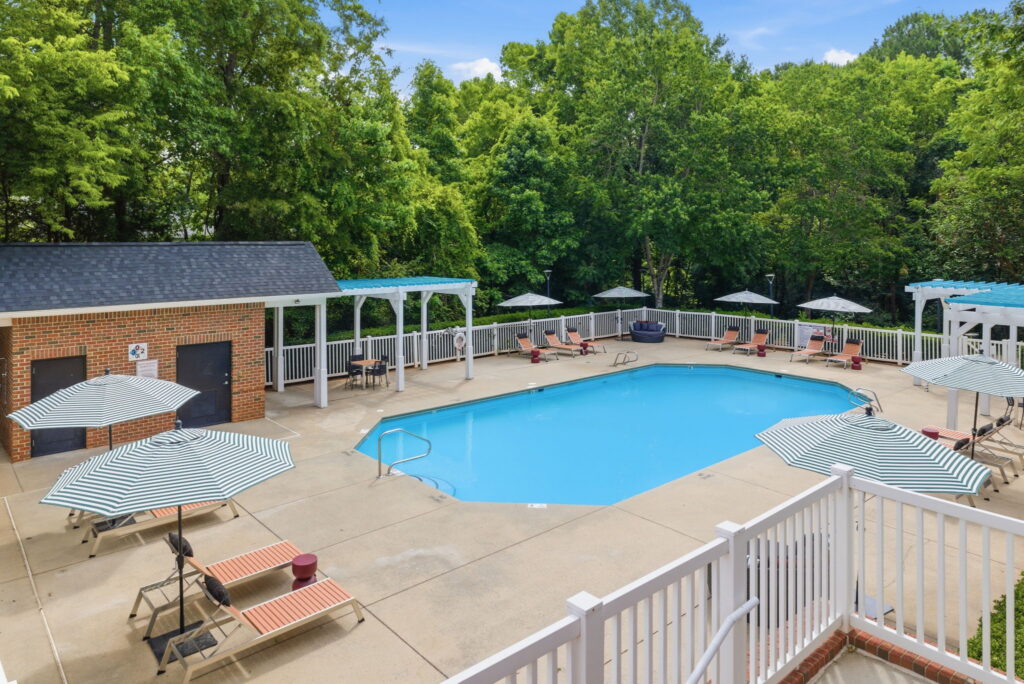 Outdoor swimming pool surrounded by lounge chairs with striped umbrellas, white fencing, and pergolas. Trees and greenery provide a natural backdrop around the pool area.